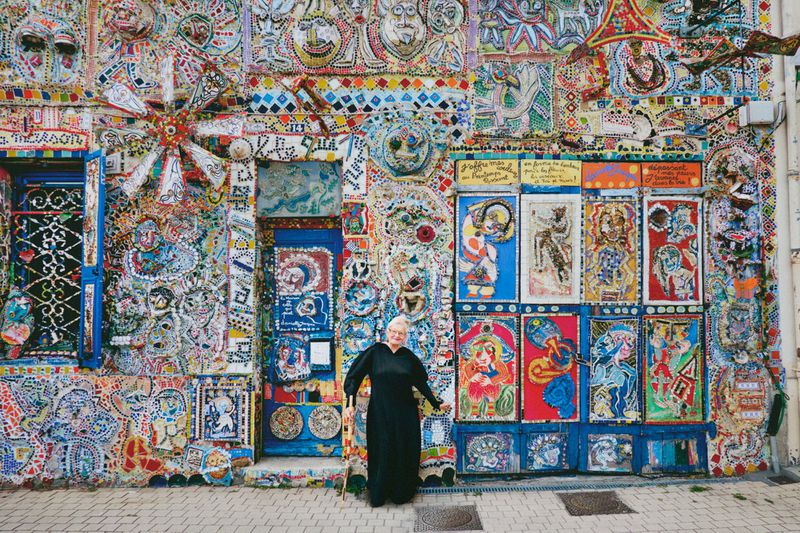 Older woman in black dress stands before a vibrantly decorated mosaic wall featuring various colorful tiles and artwork on a street.