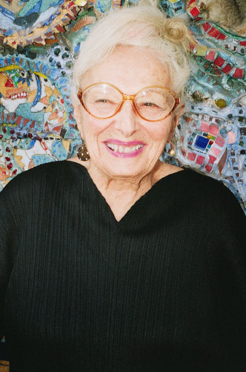 Close-up portrait of a smiling elderly woman with short white hair, wearing glasses and a black top, with a mosaic tile background.