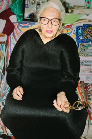 Elderly woman with glasses sits indoors wearing a black dress, holding a pair of eyeglasses in her right hand.