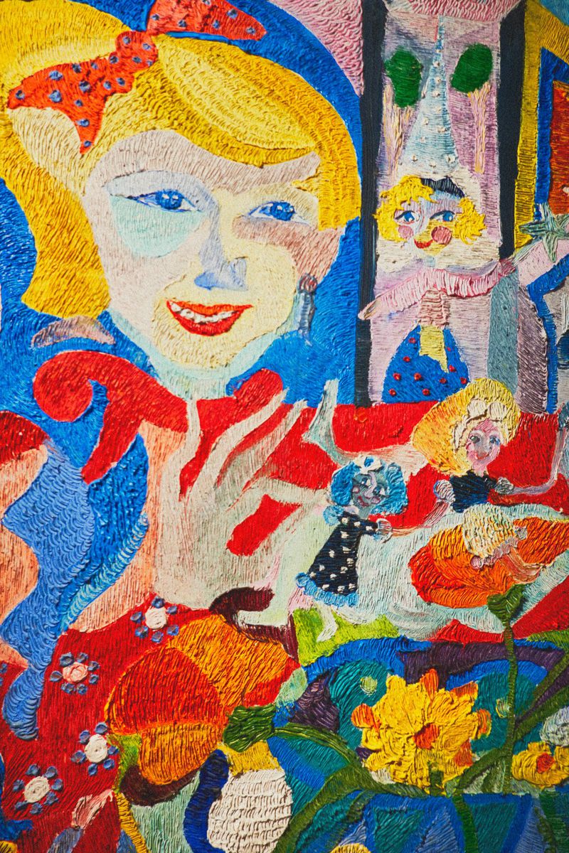 Colorful artwork of a fair-skinned girl with blond hair and a red bow, surrounded by red and yellow flowers, created with a stitching technique.