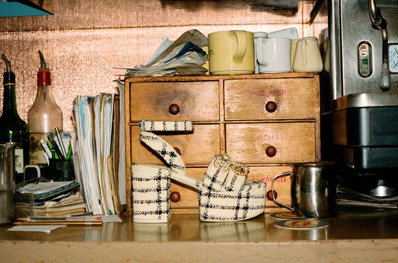 Platform heel in front of vintage wooden drawers, topped with mugs, next to an espresso maker. Bottles, pens, and papers are visible.