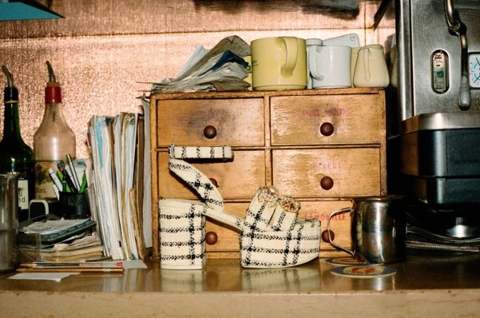 Platform heel in front of vintage wooden drawers, topped with mugs, next to an espresso maker. Bottles, pens, and papers are visible.