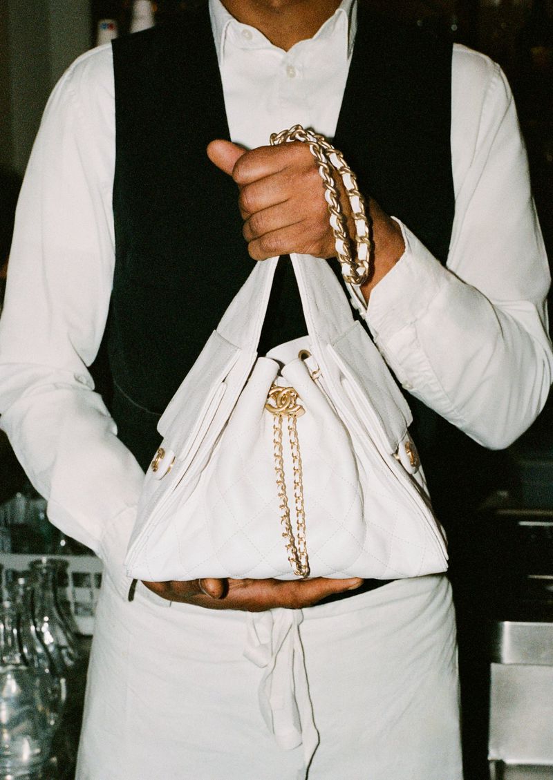 A person in a vest and shirt holding a white Chanel handbag with gold chain straps and logo, showcasing fashion and style.