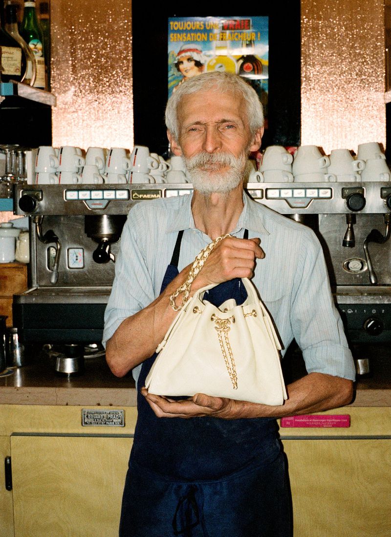Elderly man in apron holds a beige purse with gold chain detail in front of a coffee maker in a cafe setting, captured in a medium shot.