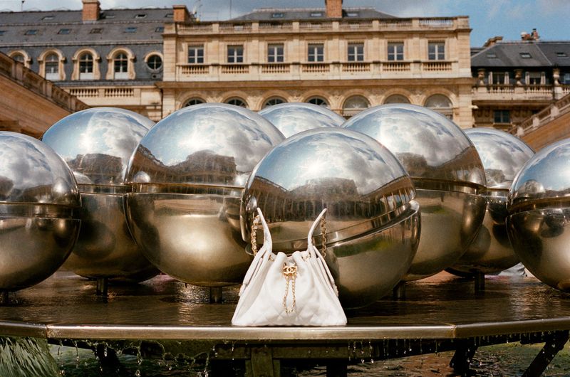 Chanel handbag positioned in front of mirrored spheres in an outdoor, urban environment with stone buildings visible in the background.
