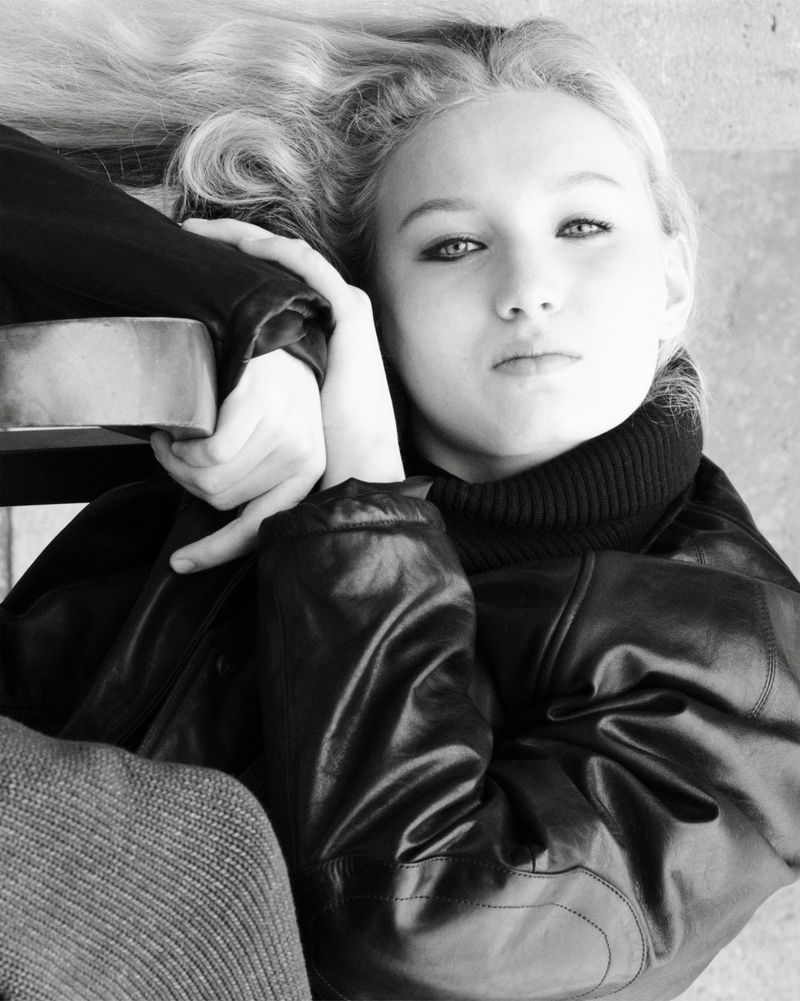 Young woman with blond hair lying down, wearing black turtleneck and leather jacket. Low-angle portrait shot in black and white.