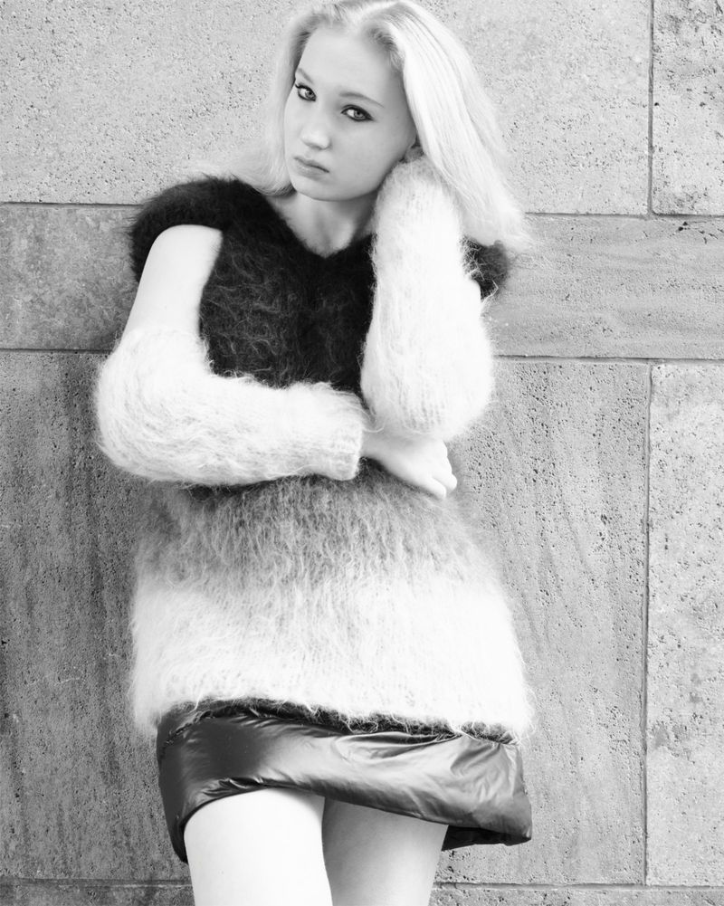 Black and white portrait of a female model in a fuzzy, short dress leaning against a stone wall, looking at the camera.