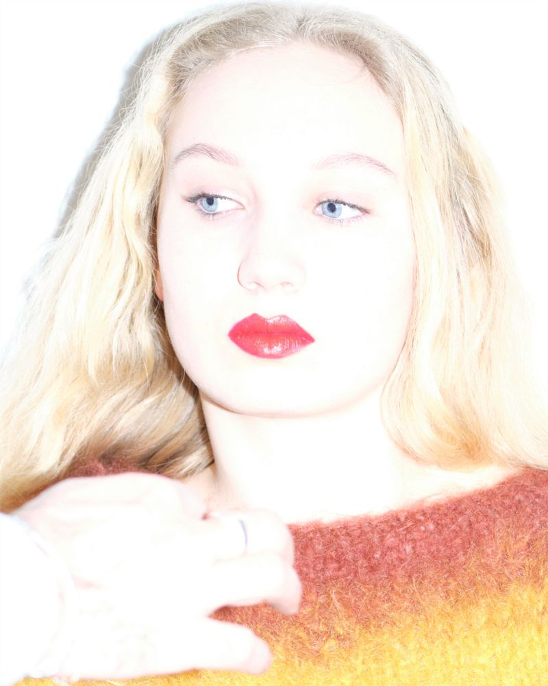 Portrait of a fair-skinned woman with long blonde hair, bright red lipstick, and a multicoloured sweater posing indoors.