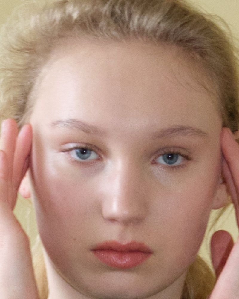Close-up portrait of a young woman with light blonde hair and blue eyes, looking directly at the camera, with her hands near her face.