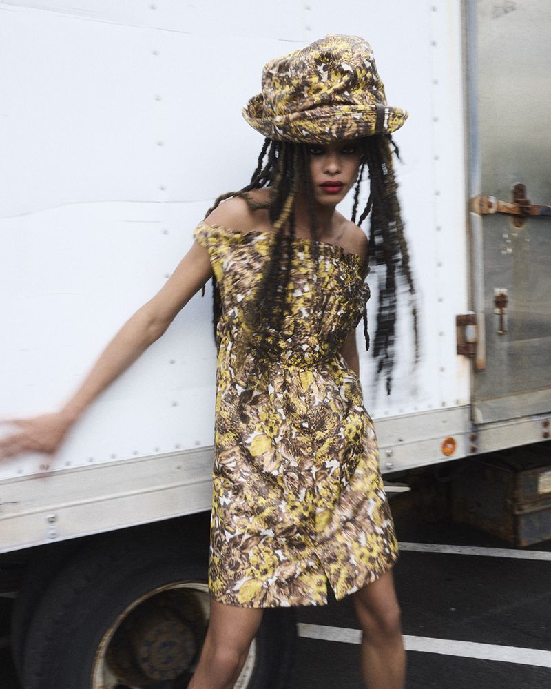 Woman poses outdoors wearing a patterned dress and bucket hat, showcasing a fashion-forward ensemble in front of a commercial vehicle.