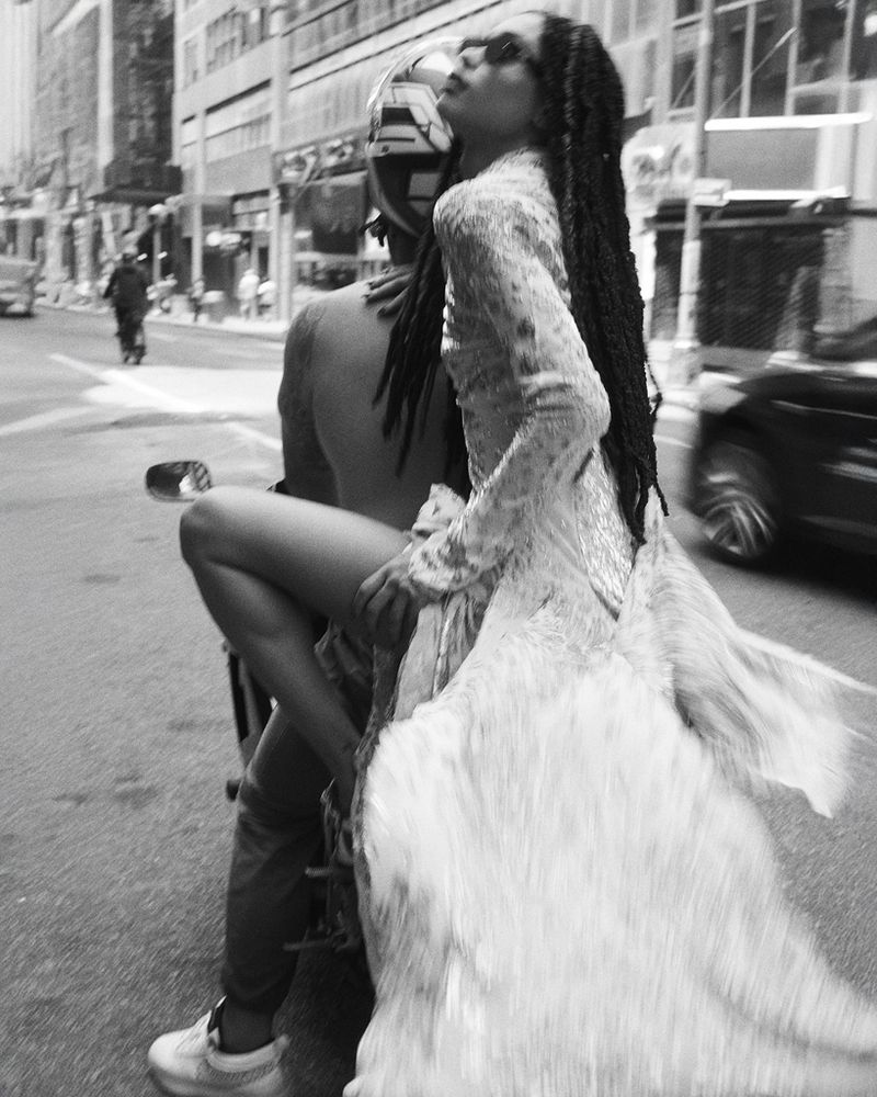 Black and white outdoor shot of a woman in a flowing dress riding a motorcycle in a city street, creating a dynamic, blurred motion effect.