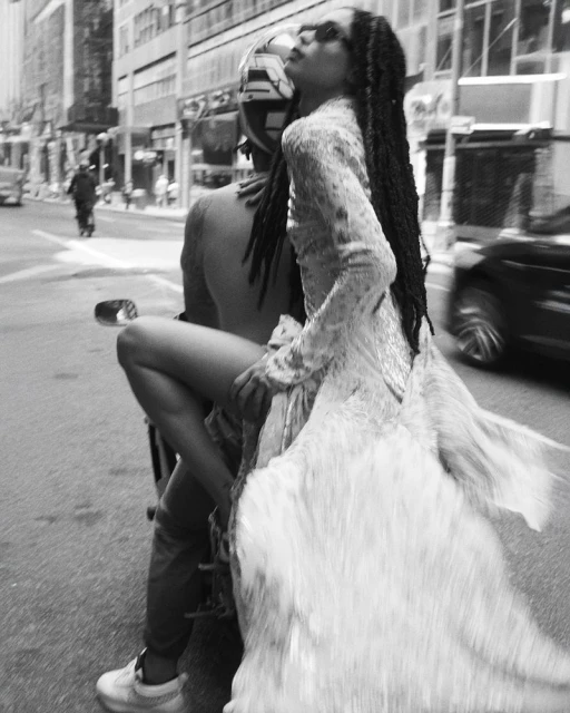 Black and white outdoor shot of a woman in a flowing dress riding a motorcycle in a city street, creating a dynamic, blurred motion effect.