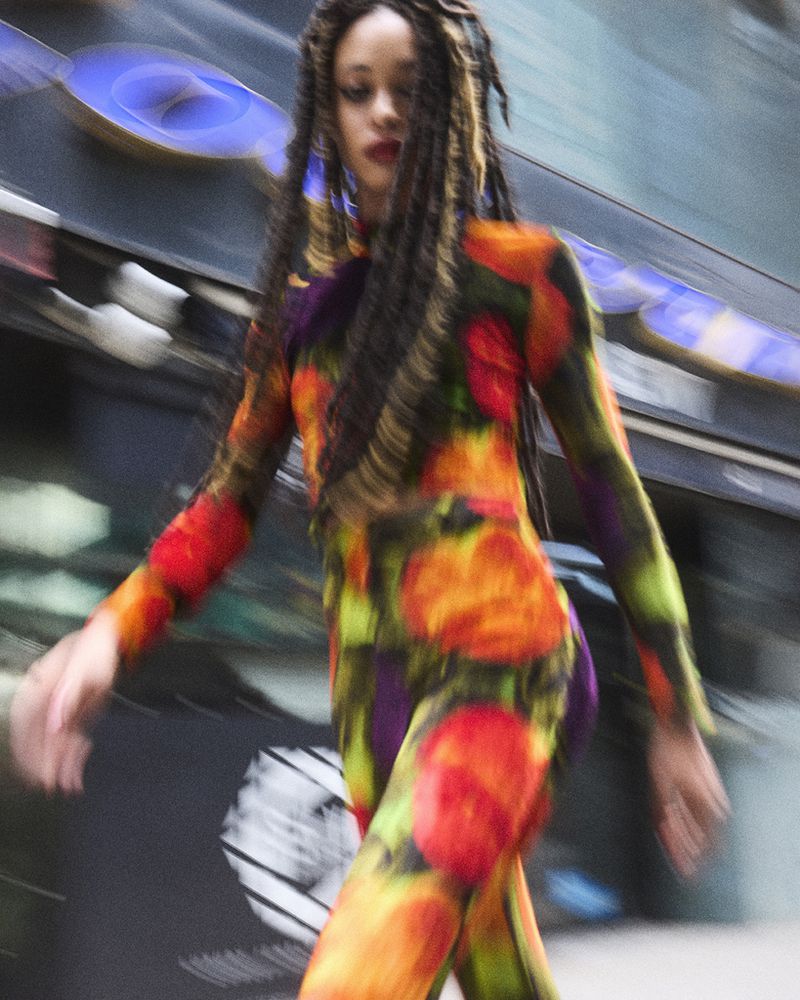 A person with long braids wearing a vibrant floral catsuit walks on a city street in blurred motion, creating a sense of movement and energy.