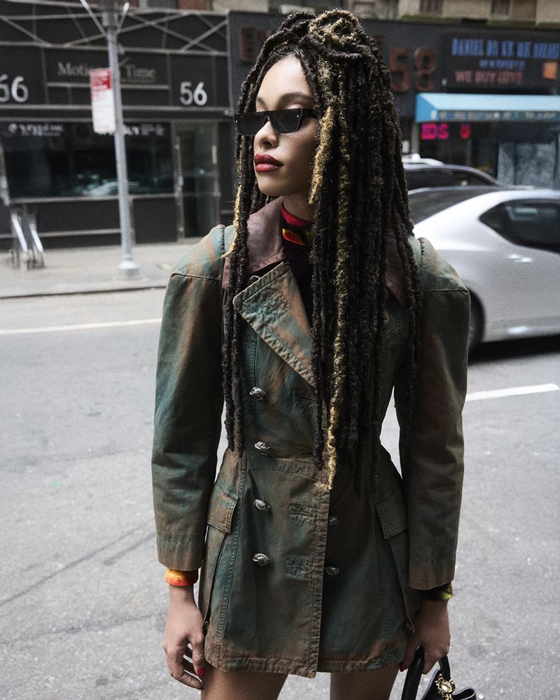 A woman with locs and sunglasses poses in an urban setting, wearing a vintage-style jacket and carrying a small bag. She's on a city street.