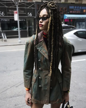 A woman with locs and sunglasses poses in an urban setting, wearing a vintage-style jacket and carrying a small bag. She's on a city street.
