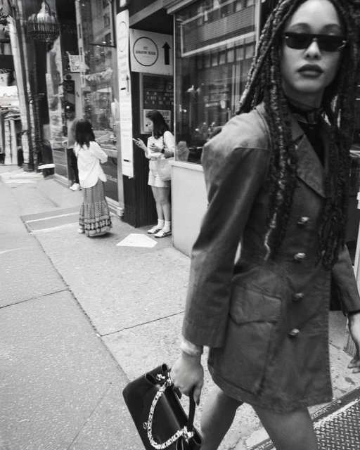 Black and white street-style fashion photograph featuring a woman in a jacket and sunglasses walking with a handbag, with other people in background.