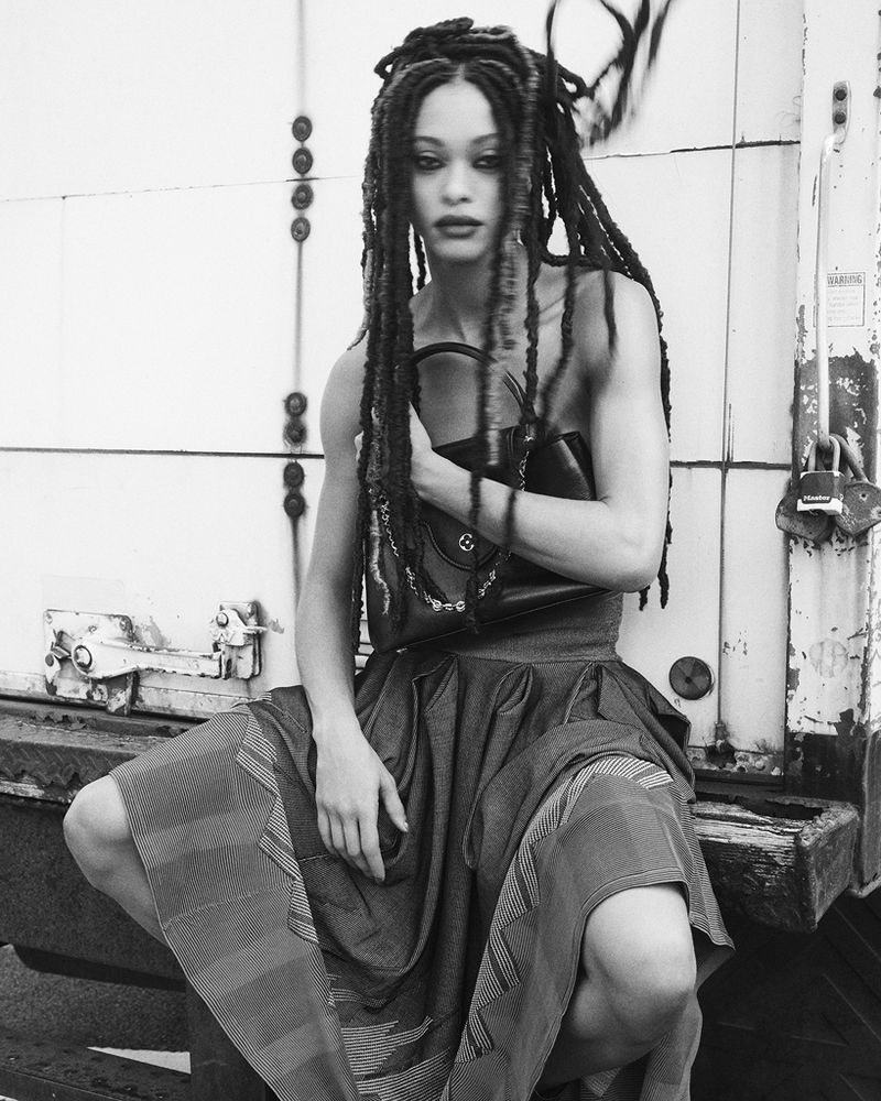 Monochrome studio shot of a fashion model with dreadlocks crouching in a patterned dress, holding a handbag against a weathered backdrop.
