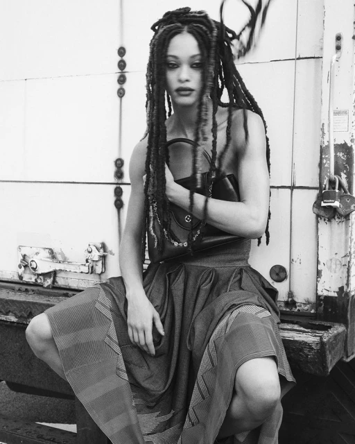 Monochrome studio shot of a fashion model with dreadlocks crouching in a patterned dress, holding a handbag against a weathered backdrop.