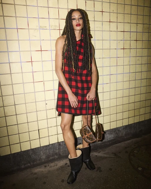 A fashionable adult woman poses in a red and black plaid dress with knee-high boots and a brown handbag against a tiled wall.