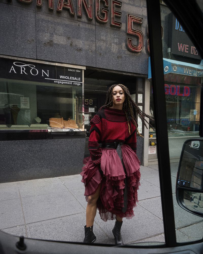 Stylish woman in a red sweater and ruffled dress poses on a city street with store fronts in the background, captured through a vehicle window.