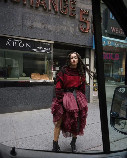 Stylish woman in a red sweater and ruffled dress poses on a city street with store fronts in the background, captured through a vehicle window.