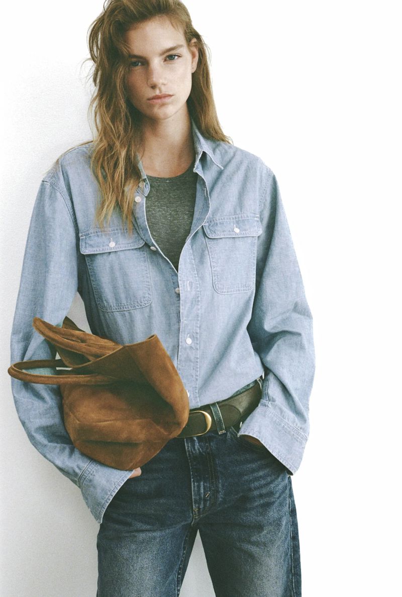 Adult woman in a relaxed pose wearing a denim button-down shirt, t-shirt, jeans, and carrying a brown leather hobo bag in studio setting.