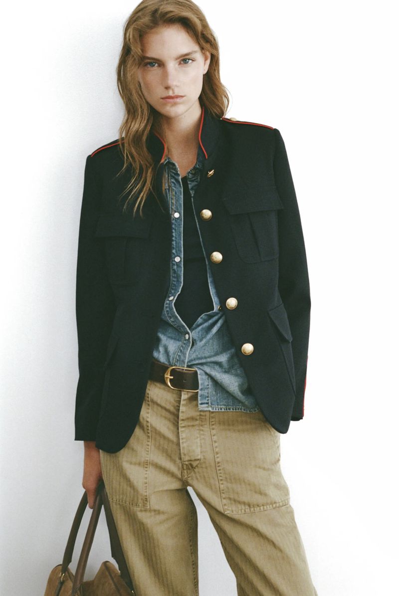 Fashionable woman in a black blazer with red trim, light blue denim shirt, and tan pants holding a brown leather tote bag, indoors.