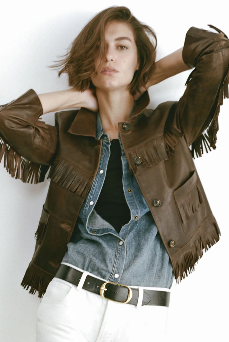 A woman poses in a brown leather fringe jacket, light blue denim shirt, black top, and white pants in a studio with white background.