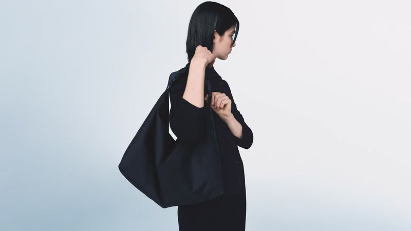 Woman in black jacket carrying a black shoulder bag against a light blue background in a fashion or product shot.