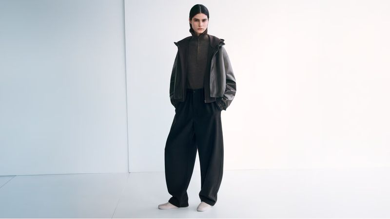 Fashion shot of a female model wearing a brown top and jacket with oversized black pants, posing in a bright, white photo studio.