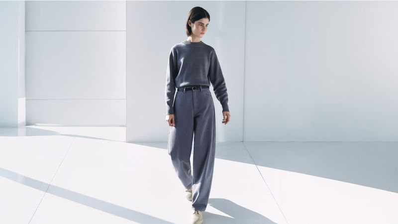 A young woman in a grey sweater and grey wide-leg pants walks in a brightly lit minimalist indoor space with neutral colors.