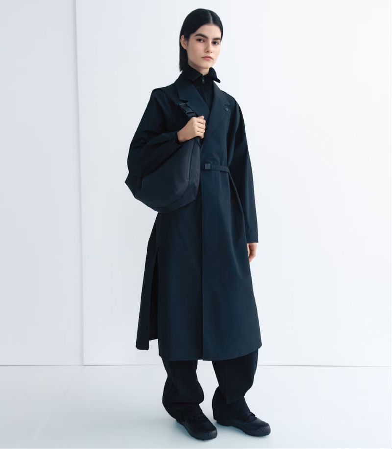 Fashion portrait of a woman in a long, black trench coat and a shoulder bag in a minimalist photo studio setting with neutral lighting.
