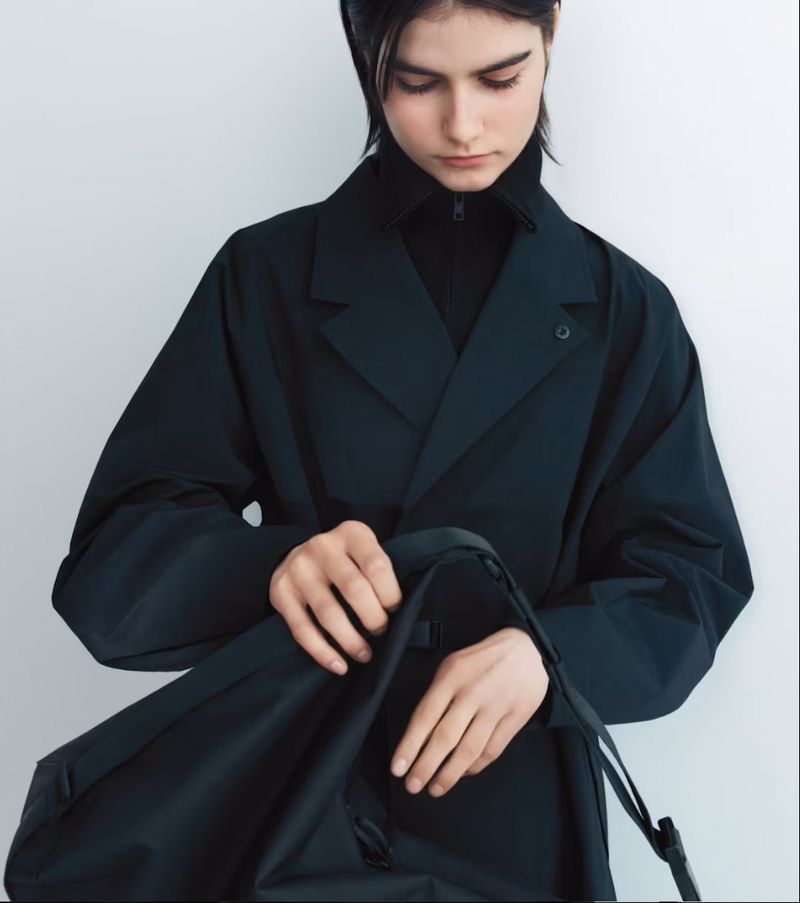 Model wearing a layered black trench jacket and opening a black bag against a bright white studio backdrop.