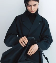 Model wearing a layered black trench jacket and opening a black bag against a bright white studio backdrop.