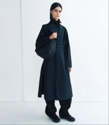 Model wearing a long black belted coat, matching wide trousers, and a shoulder bag against a white studio background.