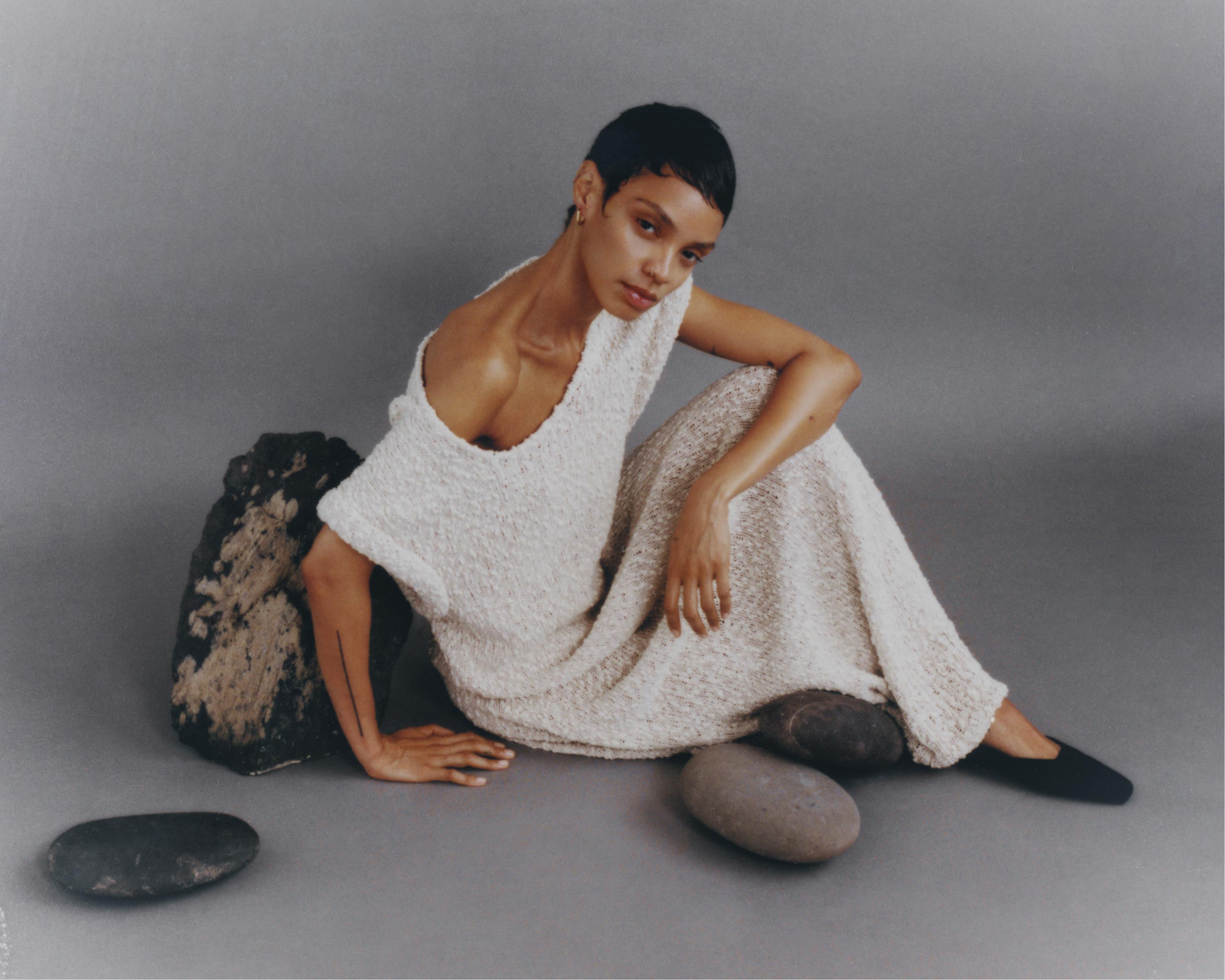 Adult woman in cream dress sits on studio floor among dark stones in a minimal fashion shot with soft studio lighting and neutral background.