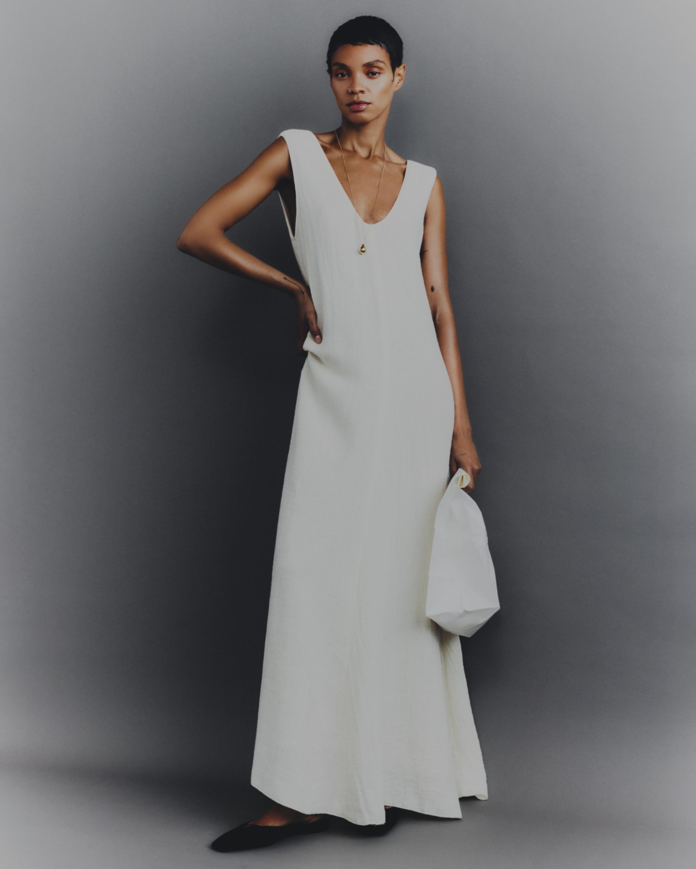 Fashion shot of a short-haired woman in an off-white sleeveless dress with a hand bag, standing in front of a grey background in a studio.