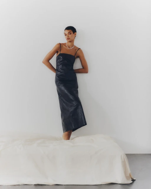 An adult woman wearing a dress and a pearl necklace poses in a studio setting near a white bed-like object.