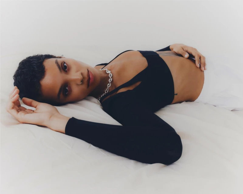 A person with dark hair and a black top lies on a white bed, looking at the camera with a neutral expression in a portrait shot.
