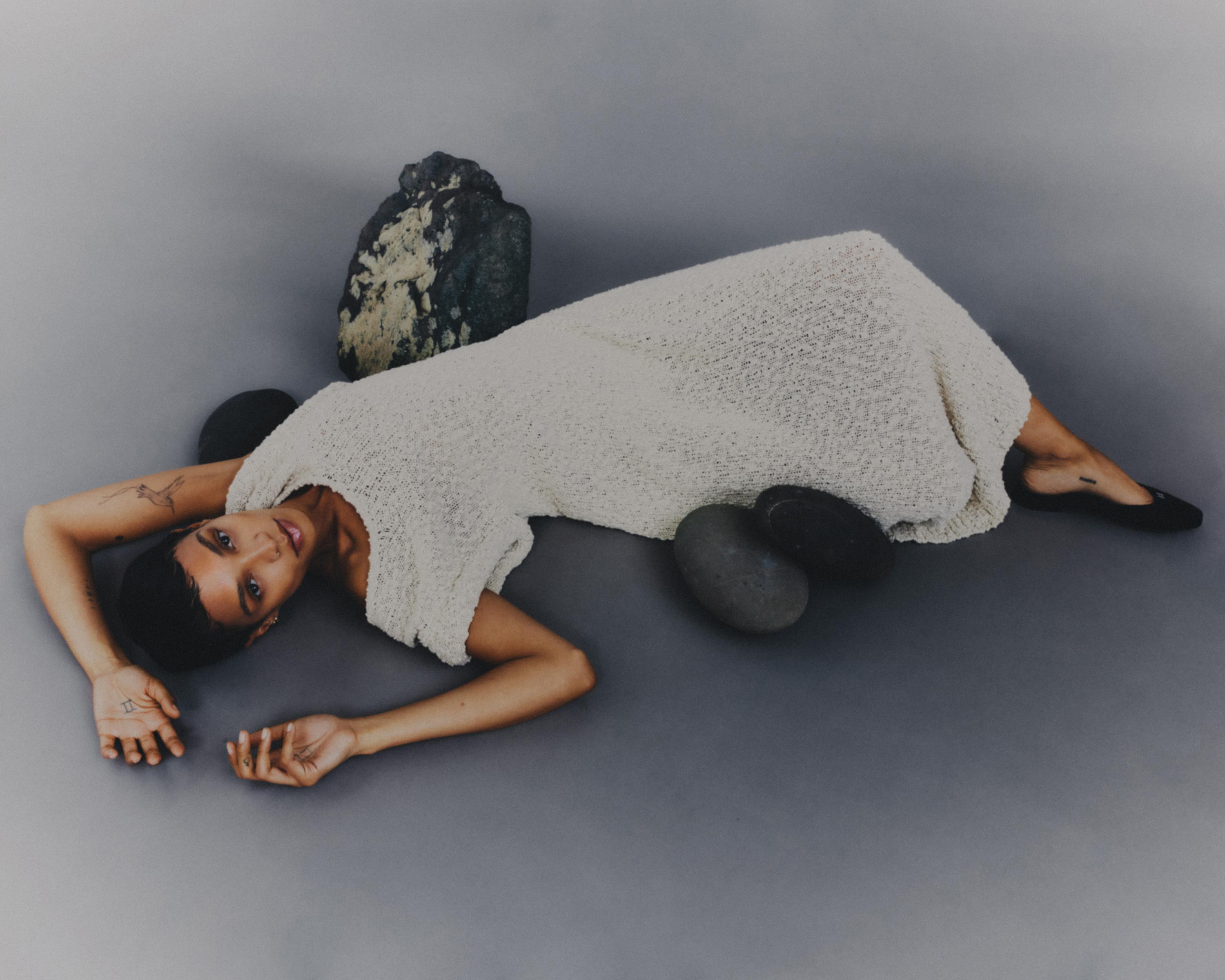 Adult woman in a cream-colored dress poses on a gray background surrounded by river stones, creating a minimalistic fashion image.
