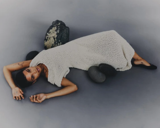 Adult woman in a cream-colored dress poses on a gray background surrounded by river stones, creating a minimalistic fashion image.