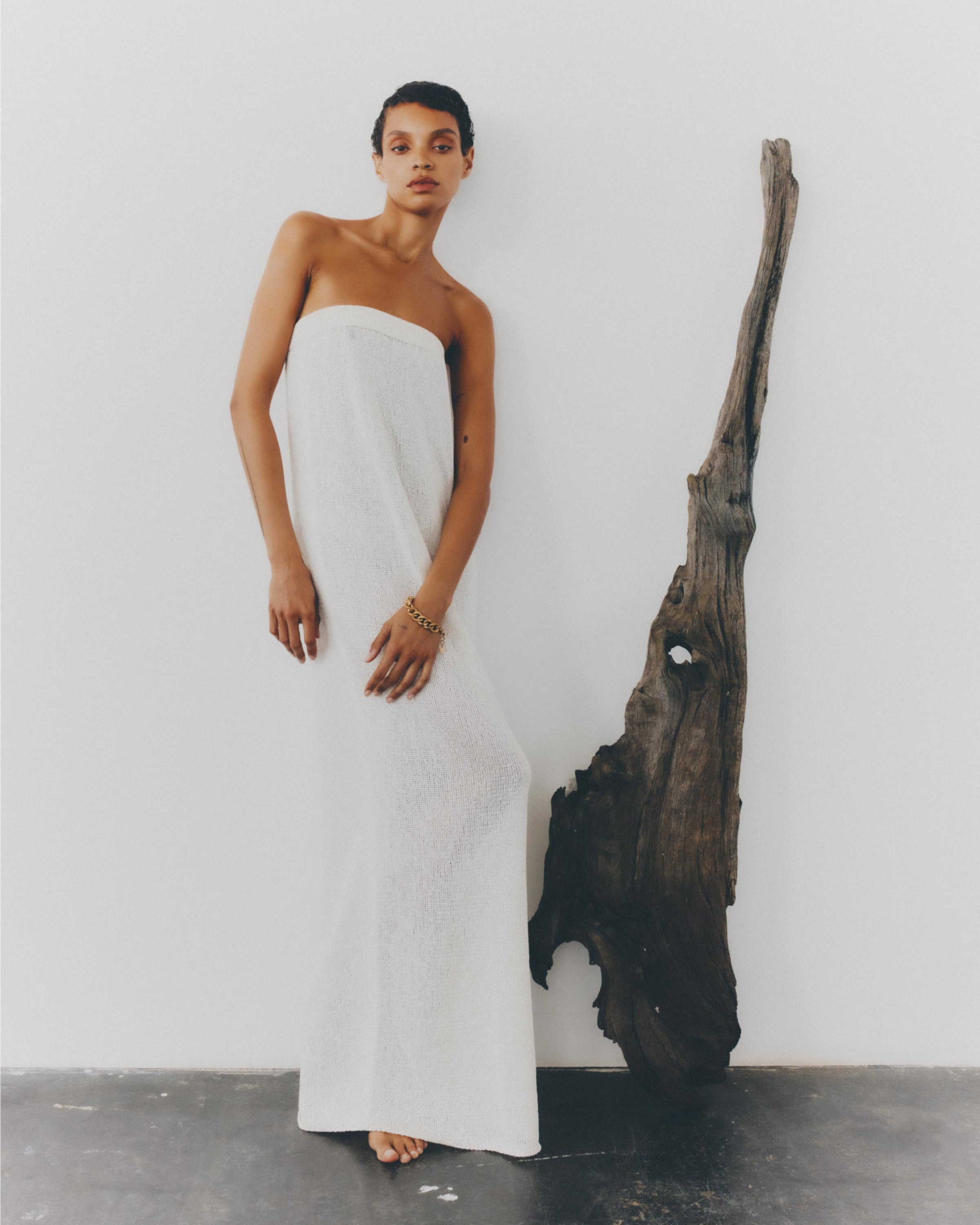 Indoor portrait shot of a slender young adult woman wearing a white dress next to a large driftwood sculpture against a white wall.