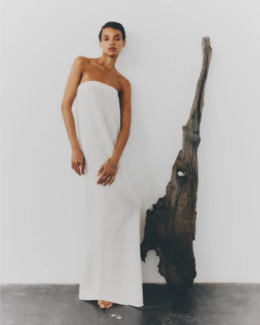 Indoor portrait shot of a slender young adult woman wearing a white dress next to a large driftwood sculpture against a white wall.