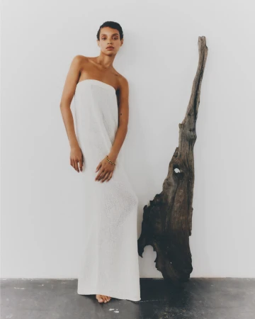 Indoor portrait shot of a slender young adult woman wearing a white dress next to a large driftwood sculpture against a white wall.