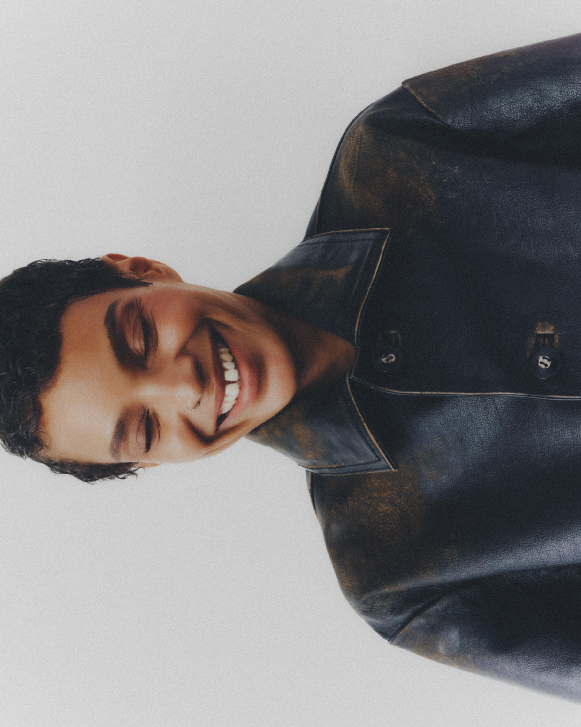 Close-up portrait of a smiling young adult with short curly hair wearing a dark leather jacket against a neutral backdrop in studio light.