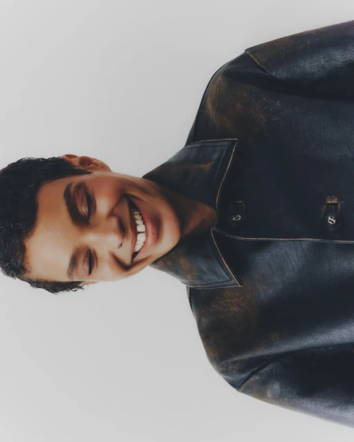 Close-up portrait of a smiling young adult with short curly hair wearing a dark leather jacket against a neutral backdrop in studio light.