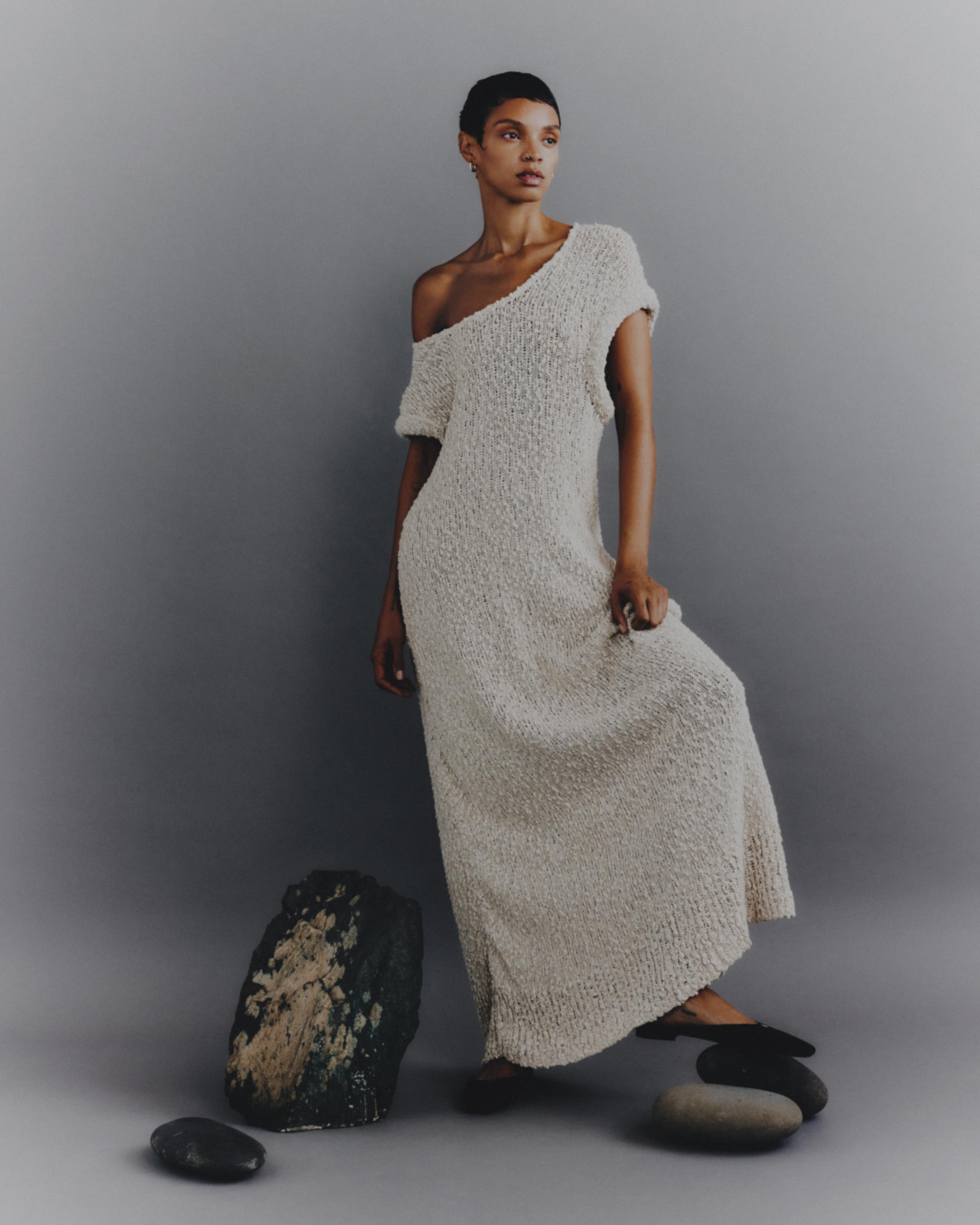 Elegant adult female poses in flowing off-shoulder knit dress indoors. Minimalist fashion portrait with neutral colors and studio setting.
