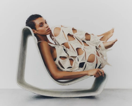 Model Akira Reid curled in a sculptural concrete rocking chair wearing an off-white cut-out dress against a white studio background.