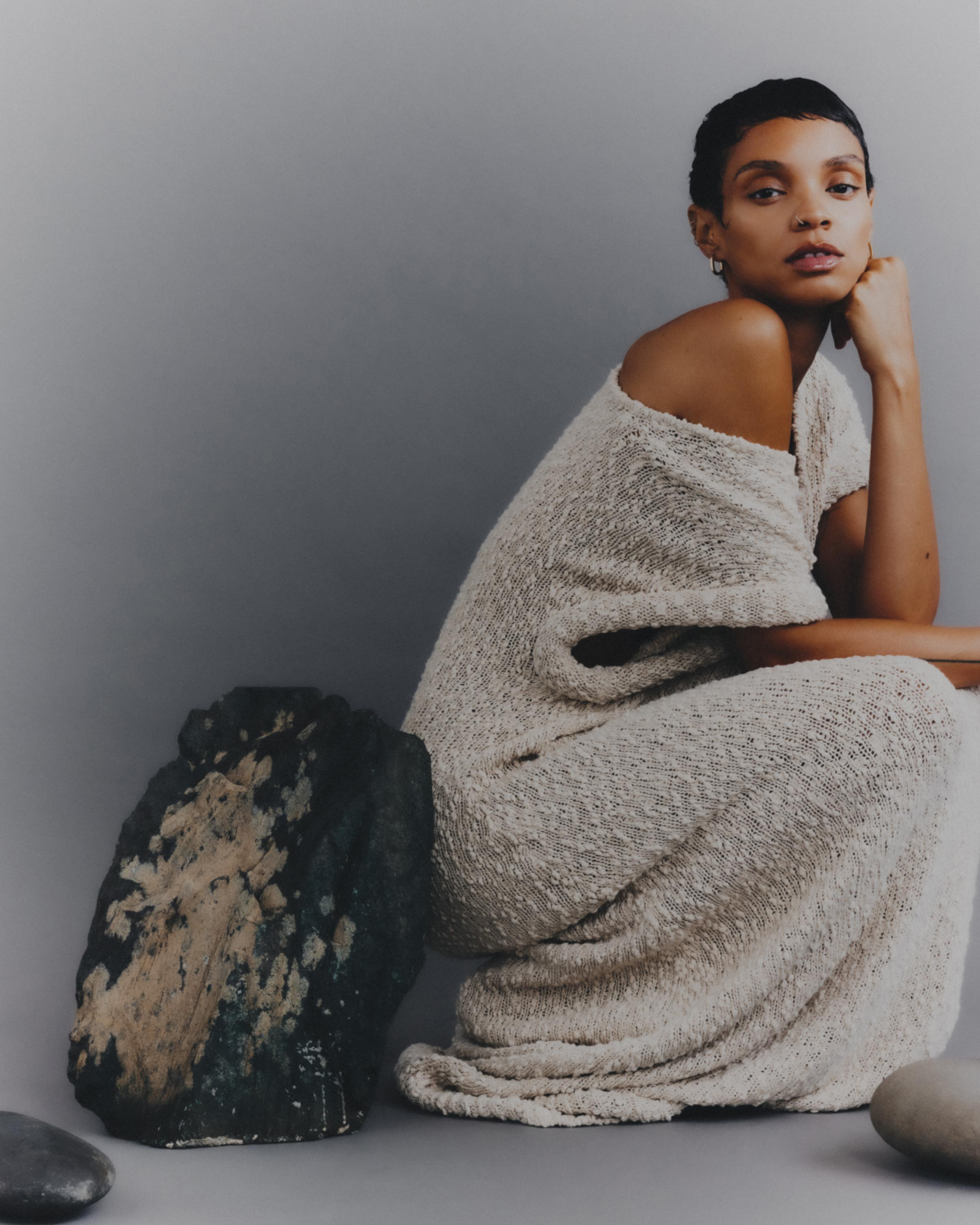 A person in a dress sitting next to a large rock against a muted background, showcasing neutral tones and minimalist styling.