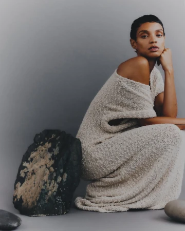 A person in a dress sitting next to a large rock against a muted background, showcasing neutral tones and minimalist styling.